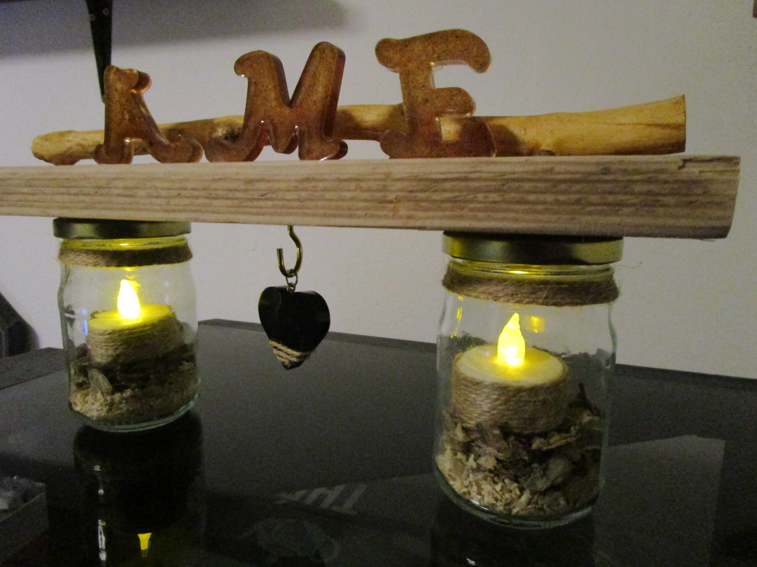 Le Murmure de l'Âme "Apportez une touche d'originalité à votre décoration avec ce luminaire DIY. Deux bocaux en verre, transformés en bougeoirs, sont reliés par une structure en bois brut. Des lettres en bois, formant le mot "AME", apportent une touche décorative et personnalisée. Un cœur en bois suspendu complète cette création unique."