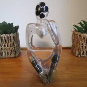 Penseur de cristal Façonnée à la main, cette pièce unique révèle une silhouette épurée, symbole universel de la pensée et de la méditation. La résine cristalline, tel un écrin transparent, laisse entrevoir un trésor de pierres naturelles : des perles de tourmaline, noires et mystérieuses, qui captent la lumière et diffusent une énergie protectrice. Des éclats d'argent, tels des fragments d'étoiles, scintillent au cœur de la matière, apportant une touche de lumière et de raffinement.
