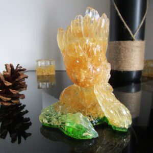 Sculpture de collection "Groot du Bois" faite à la main, en résine transparente et copeaux de bois. La base est en résine verte, évoquant la nature. On voit Groot de face, avec une texture lumineuse et naturelle. Des pommes de pin sont disposées à proximité.