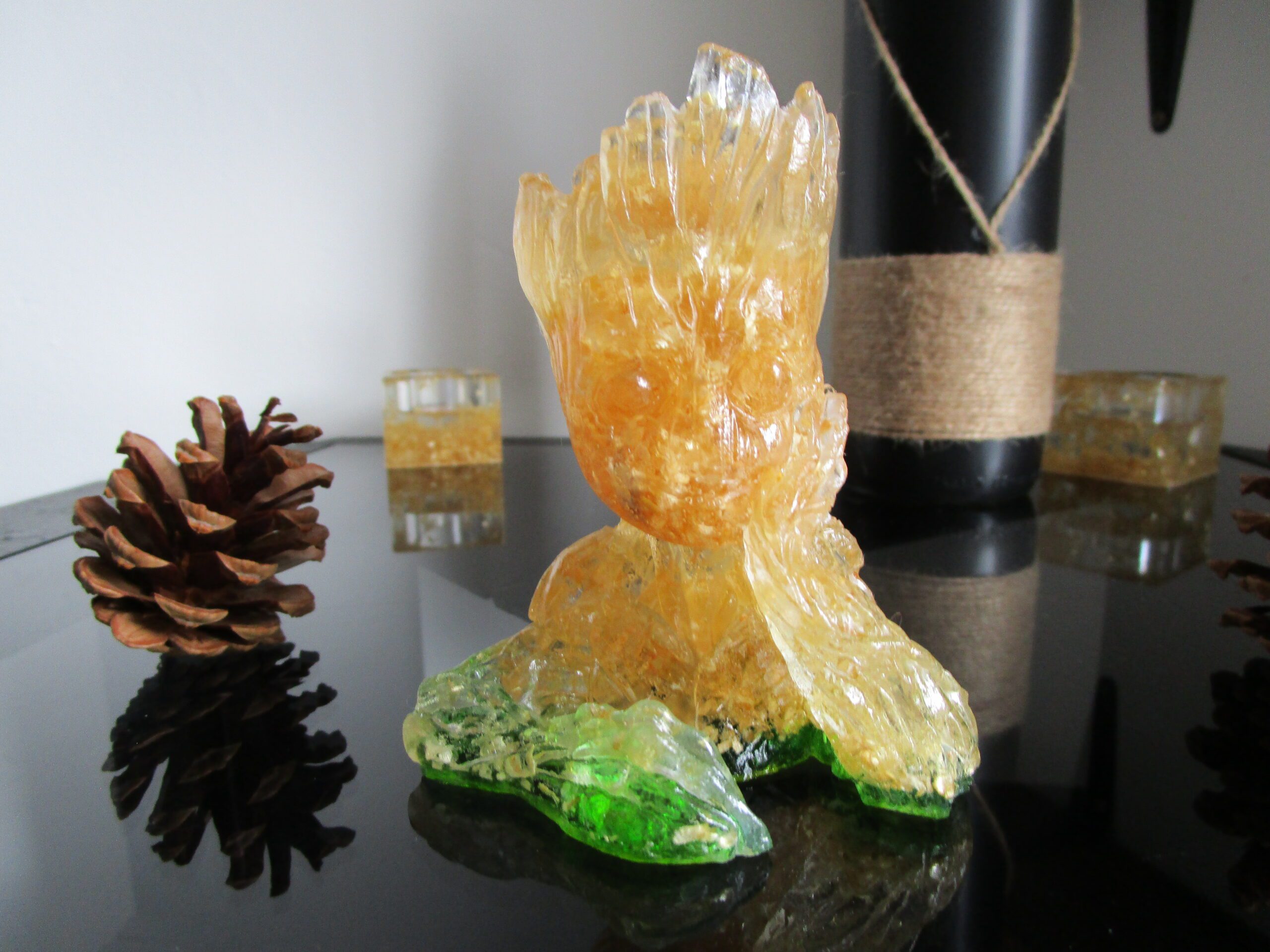 Sculpture de collection "Groot du Bois" faite à la main, en résine transparente et copeaux de bois. La base est en résine verte, évoquant la nature. On voit Groot de face, avec une texture lumineuse et naturelle. Des pommes de pin sont disposées à proximité.