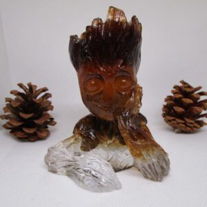 Sculpture de Groot Sciure , faite à la main, en résine et sciure de bois, avec des pierres naturelles incrustées. On voit Groot de face, avec un visage souriant et des yeux ronds. Ses bras sont posés sur une base transparente. Des pommes de pin sont disposées de chaque côté de la sculpture.