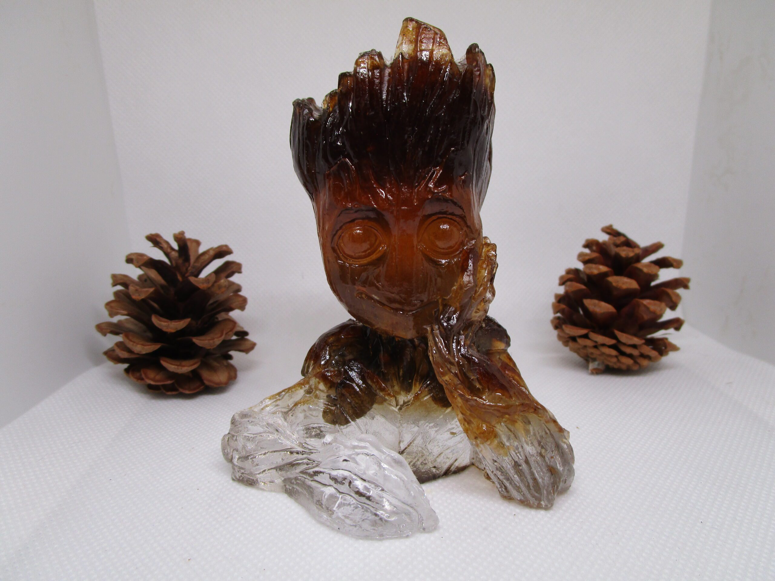 Sculpture de Groot Sciure , faite à la main, en résine et sciure de bois, avec des pierres naturelles incrustées. On voit Groot de face, avec un visage souriant et des yeux ronds. Ses bras sont posés sur une base transparente. Des pommes de pin sont disposées de chaque côté de la sculpture.