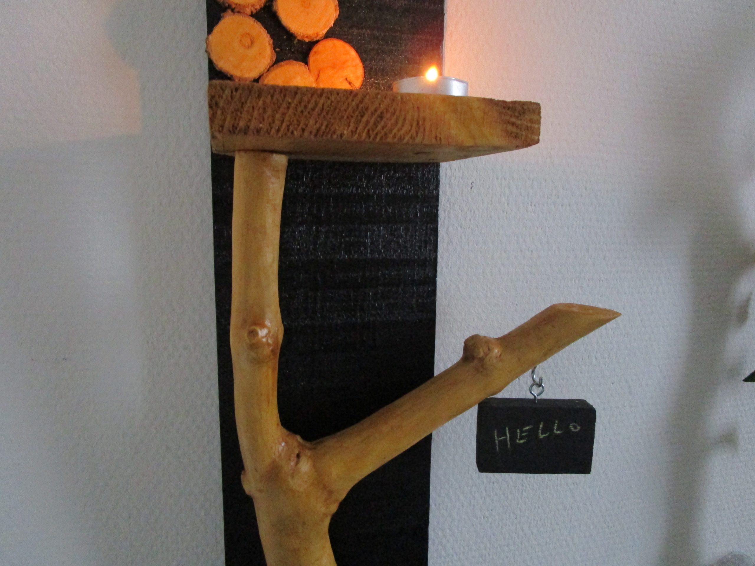 Une branche d'arbre, aux formes organiques et irrégulières, s'élève depuis la base. Elle soutient une petite étagère en bois clair, où repose une bougie chauffe-plat. La flamme vacillante crée une ambiance chaleureuse et intime. Un petit morceau de bois, peint en ardoise, est suspendu à l'extrémité de la branche. Vous pouvez y écrire un mot doux, un message inspirant ou simplement votre prénom. Cette ardoise personnalisable ajoute une touche ludique et interactive à la création.