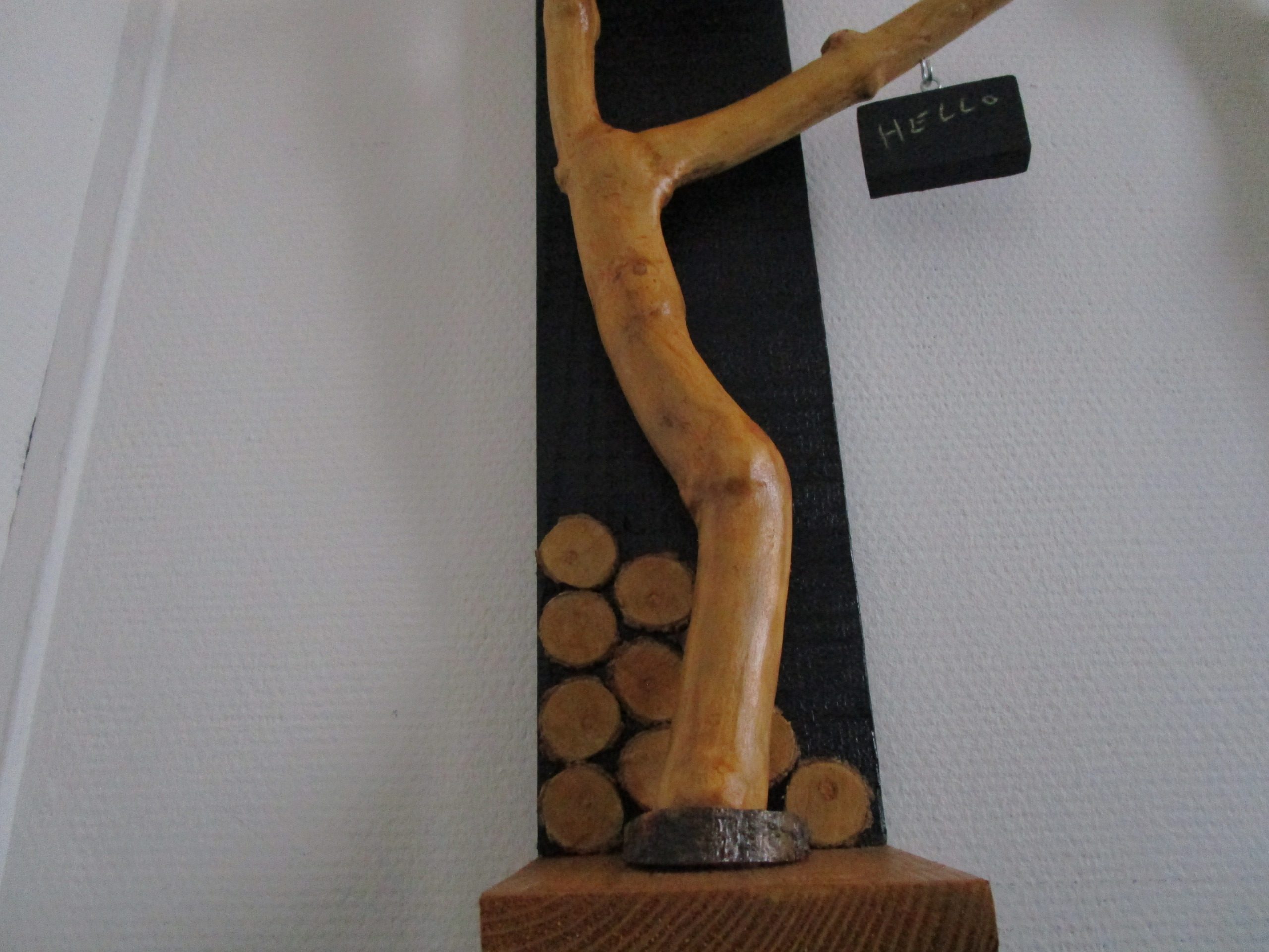 Une branche d'arbre, aux formes organiques et irrégulières, s'élève depuis la base. Elle soutient une petite étagère en bois clair, où repose une bougie chauffe-plat. La flamme vacillante crée une ambiance chaleureuse et intime. Un petit morceau de bois, peint en ardoise, est suspendu à l'extrémité de la branche. Vous pouvez y écrire un mot doux, un message inspirant ou simplement votre prénom. Cette ardoise personnalisable ajoute une touche ludique et interactive à la création.