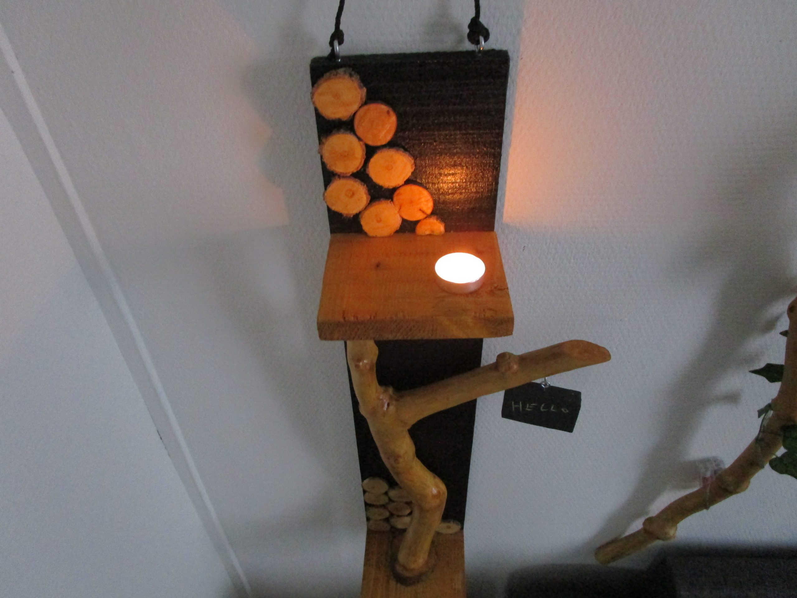 Une branche d'arbre, aux formes organiques et irrégulières, s'élève depuis la base. Elle soutient une petite étagère en bois clair, où repose une bougie chauffe-plat. La flamme vacillante crée une ambiance chaleureuse et intime. Un petit morceau de bois, peint en ardoise, est suspendu à l'extrémité de la branche. Vous pouvez y écrire un mot doux, un message inspirant ou simplement votre prénom. Cette ardoise personnalisable ajoute une touche ludique et interactive à la création.