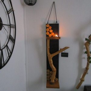 La Mémoire de l'Arbre Une branche d'arbre, aux formes organiques et irrégulières, s'élève depuis la base. Elle soutient une petite étagère en bois clair, où repose une bougie chauffe-plat. La flamme vacillante crée une ambiance chaleureuse et intime. Un petit morceau de bois, peint en ardoise, est suspendu à l'extrémité de la branche. Vous pouvez y écrire un mot doux, un message inspirant ou simplement votre prénom. Cette ardoise personnalisable ajoute une touche ludique et interactive à la création.