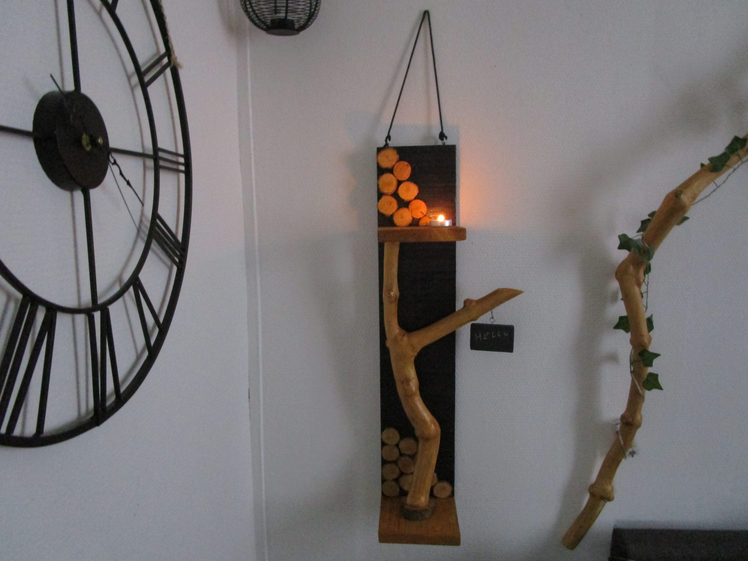 La Mémoire de l'Arbre Une branche d'arbre, aux formes organiques et irrégulières, s'élève depuis la base. Elle soutient une petite étagère en bois clair, où repose une bougie chauffe-plat. La flamme vacillante crée une ambiance chaleureuse et intime. Un petit morceau de bois, peint en ardoise, est suspendu à l'extrémité de la branche. Vous pouvez y écrire un mot doux, un message inspirant ou simplement votre prénom. Cette ardoise personnalisable ajoute une touche ludique et interactive à la création.