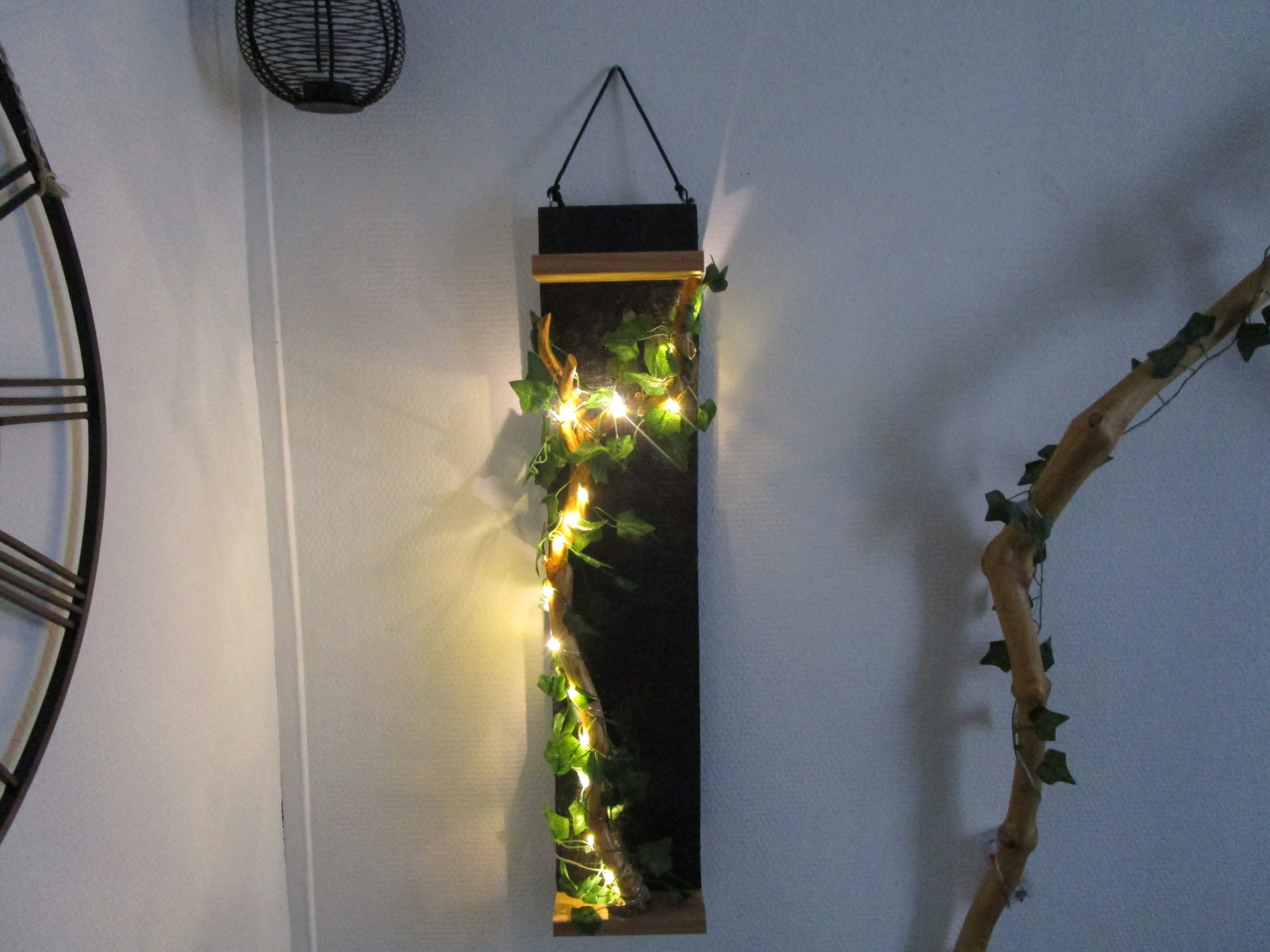 "Découvrez cette création murale unique, un mariage subtil de design moderne et de touches naturelles. Une structure rectangulaire épurée, soulignée par des finitions en bois clair, sert de support à une guirlande lumineuse LED. Celle-ci, enlacée de feuilles artificielles, crée un contraste saisissant entre la sophistication de la forme et la douceur organique de la végétation. Cette pièce apportera une touche de lumière et de verdure à votre intérieur, tout en conservant une esthétique minimaliste et contemporaine."