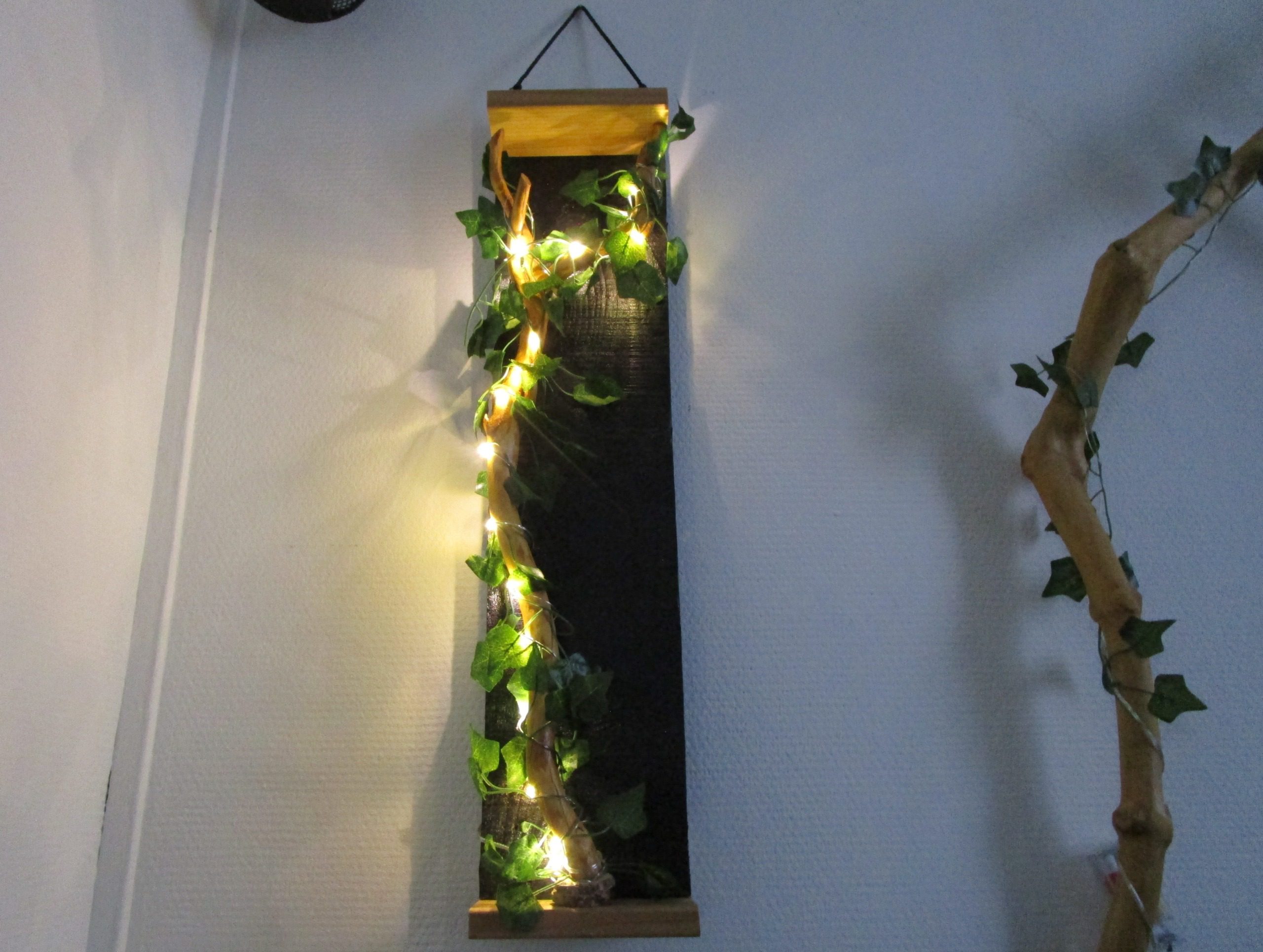 "Découvrez cette création murale unique, un mariage subtil de design moderne et de touches naturelles. Une structure rectangulaire épurée, soulignée par des finitions en bois clair, sert de support à une guirlande lumineuse LED. Celle-ci, enlacée de feuilles artificielles, crée un contraste saisissant entre la sophistication de la forme et la douceur organique de la végétation. Cette pièce apportera une touche de lumière et de verdure à votre intérieur, tout en conservant une esthétique minimaliste et contemporaine."