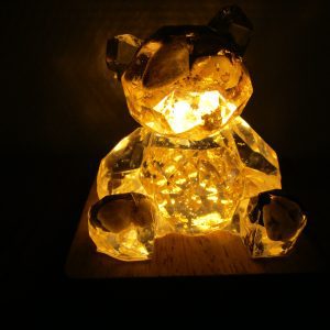 "Image d'une sculpture d'ours en résine transparente, incrusté de pierres naturelles illuminée de l'intérieur par une lumière jaune chaude. L'ours a un design géométrique moderne, avec des facettes qui captent et diffusent la lumière."