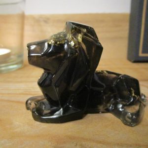 Sculpture en résine d'un lion. Fait avec passion, ce gardien des ténèbres révèle un cœur d'or pur. Des fragments de feuilles d'or, tels des éclats d'étoiles filantes, illuminent sa silhouette ténébreuse, créant un contraste saisissant entre l'obscurité et la lumière
