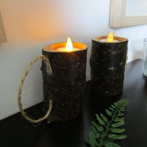 Flammes Sylvestres. Deux bougeoirs artisanaux en bois, avec des bougies allumées, posés sur une surface brillante et sombre. Les bougeoirs sont faits de morceaux de bois avec l'écorce préservée, et chacun contient une bougie blanche allumée.