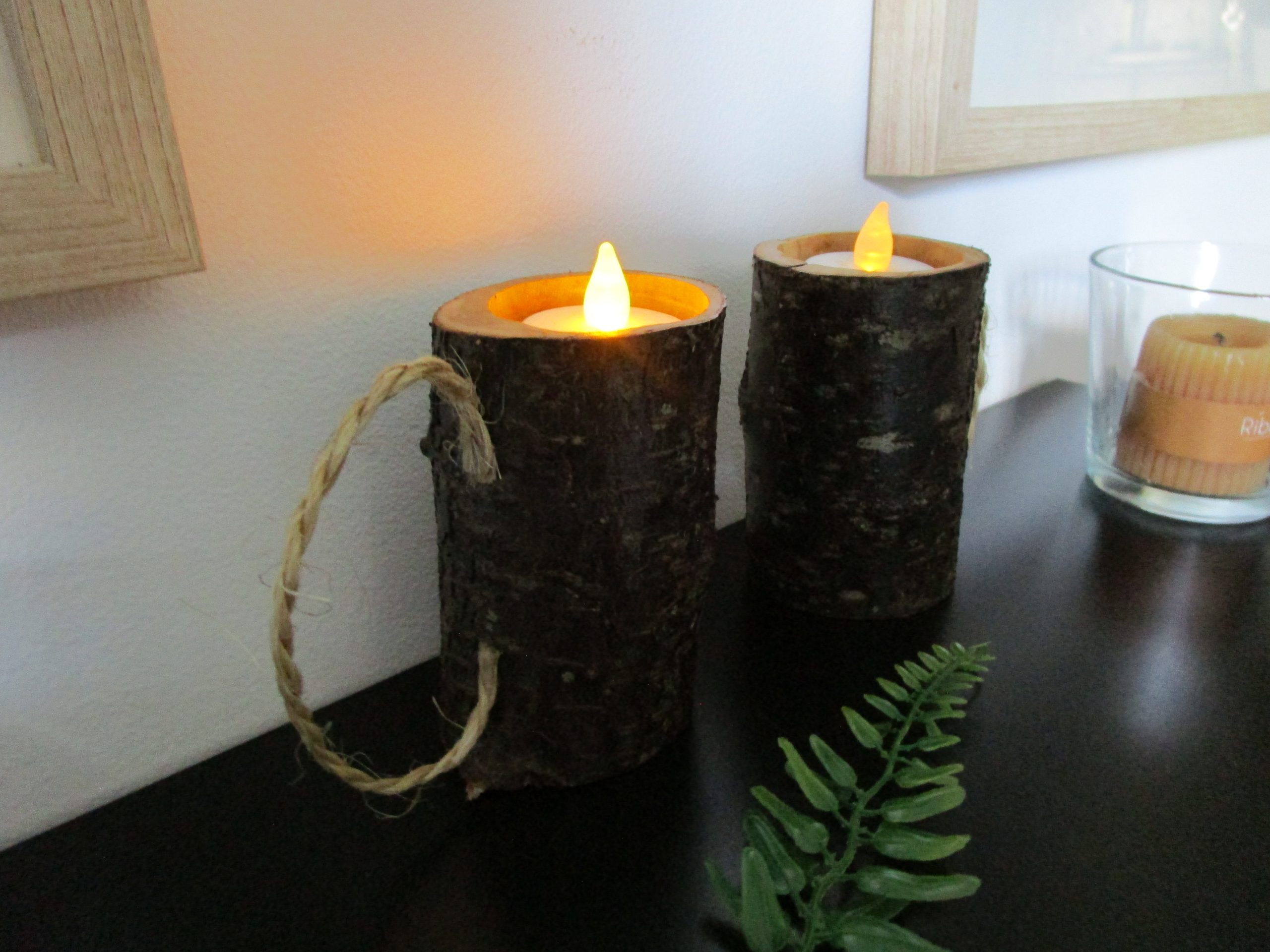 Flammes Sylvestres. Deux bougeoirs artisanaux en bois, avec des bougies allumées, posés sur une surface brillante et sombre. Les bougeoirs sont faits de morceaux de bois avec l'écorce préservée, et chacun contient une bougie blanche allumée.