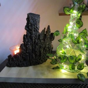Lampe décorative faite main "Rêverie Lumineuse d'une Nuit d'Été". Elle comprend une base en bois clair, une bouteille en verre entourée d'une guirlande lumineuse et de feuilles vertes, un morceau d'écorce sombre à l'arrière et un bougeoir en verre avec une bougie allumée.
