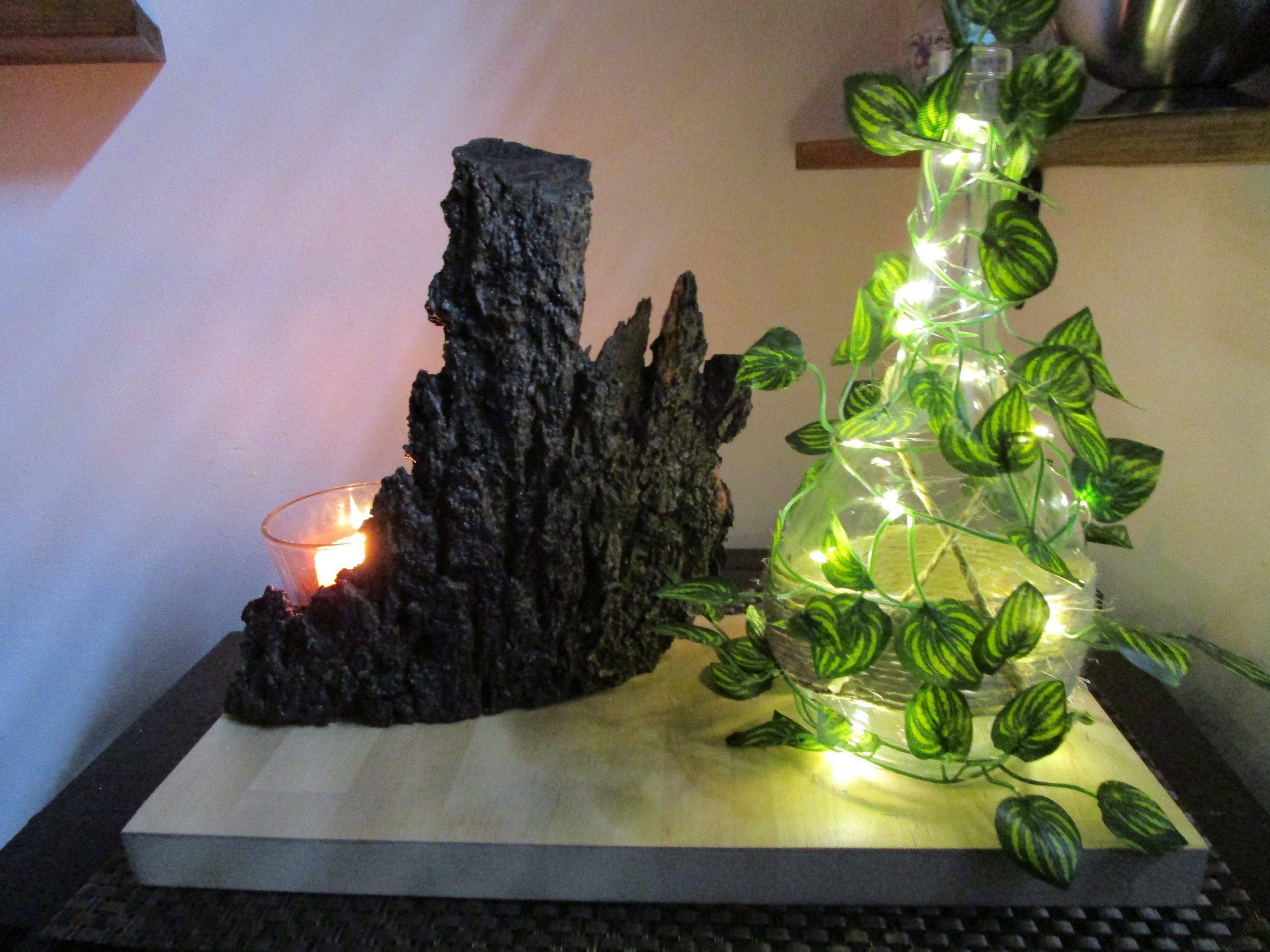 Lampe décorative faite main "Rêverie Lumineuse d'une Nuit d'Été". Elle comprend une base en bois clair, une bouteille en verre entourée d'une guirlande lumineuse et de feuilles vertes, un morceau d'écorce sombre à l'arrière et un bougeoir en verre avec une bougie allumée.