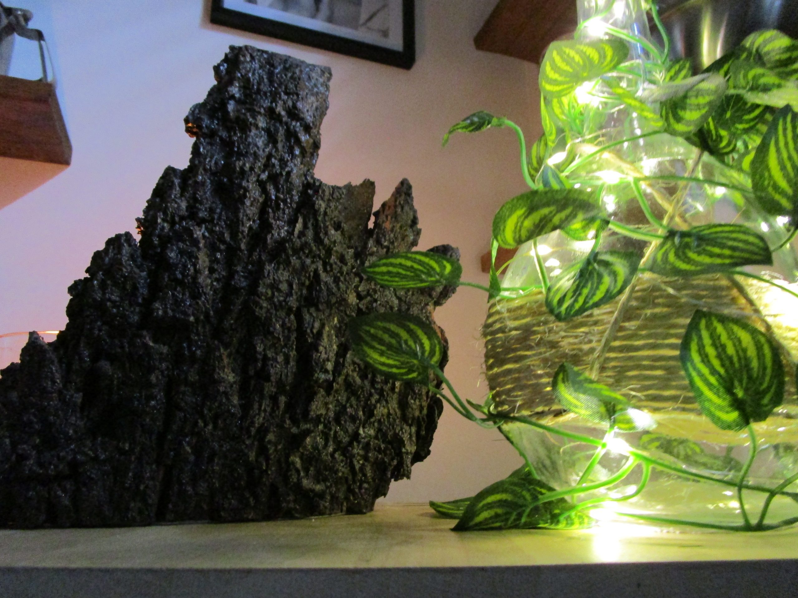 Lampe décorative faite main "Rêverie Lumineuse d'une Nuit d'Été". Elle comprend une base en bois clair, une bouteille en verre entourée d'une guirlande lumineuse et de feuilles vertes, un morceau d'écorce sombre à l'arrière et un bougeoir en verre avec une bougie allumée.
