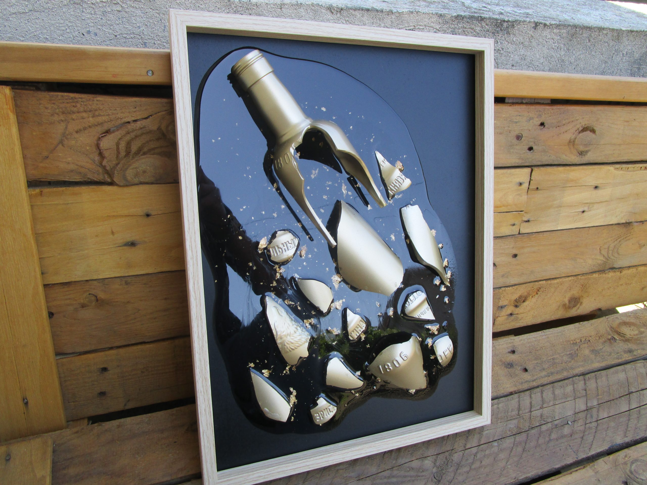 Un cadre en résine transparente incrusté d'une bouteille de vodka Eristoff dorée brisée et de feuilles d'or, avec un fond noir peint.