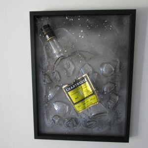 Cadre décoratif "Père Chartreux" représentant une bouteille de Chartreuse brisée et incrustée dans de la résine transparente avec des feuilles d'argent, sur un fond gris aux reflets noirs.