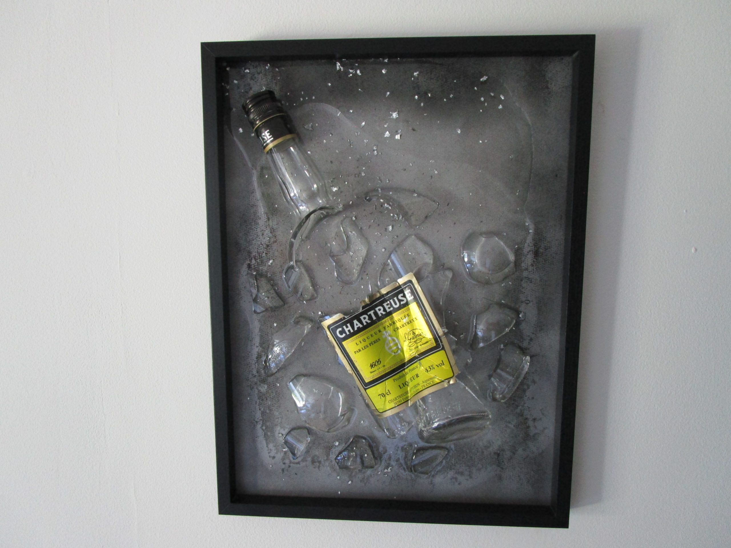 Cadre décoratif "Père Chartreux" représentant une bouteille de Chartreuse brisée et incrustée dans de la résine transparente avec des feuilles d'argent, sur un fond gris aux reflets noirs.