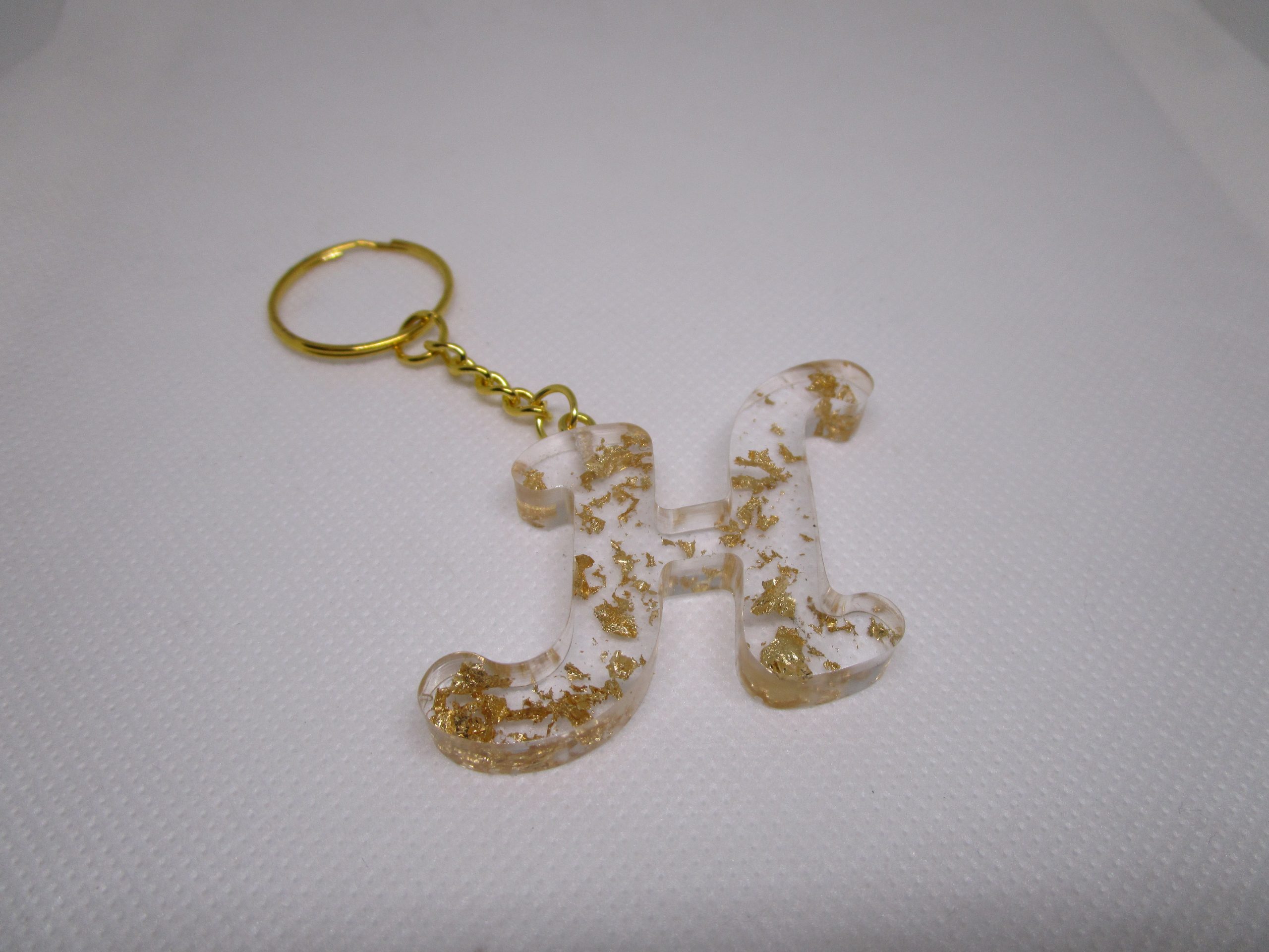 Porte-clés H : une lettre d'or capturée. La résine cristalline murmure des secrets. Des paillettes dorées dansent. Un bijou personnel, lumineux. Il chante votre initiale. Un éclat discret.