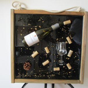 L'Écrin du Vignoble Cadre artistique mural avec une bouteille de vin verte, des bouchons de liège, un verre et une pomme de pin, le tout figé dans de la résine transparente sur un fond noir avec des paillettes dorées, encadré de bois vieilli.
