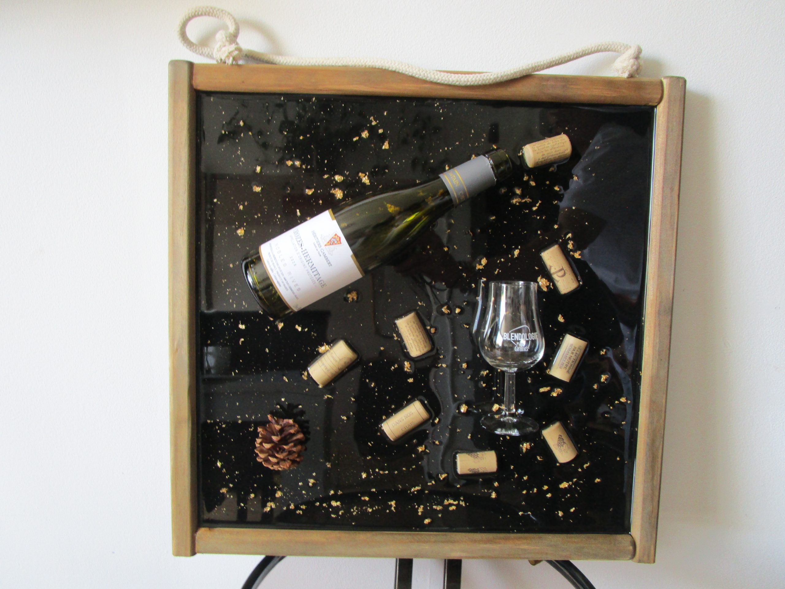 L'Écrin du Vignoble Cadre artistique mural avec une bouteille de vin verte, des bouchons de liège, un verre et une pomme de pin, le tout figé dans de la résine transparente sur un fond noir avec des paillettes dorées, encadré de bois vieilli.