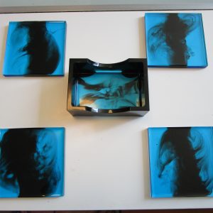 Blue Service. Quatre dessous de verre carrés et un socle de rangement rectangulaire, en résine bleu lagon et noir, posés sur une table blanche.