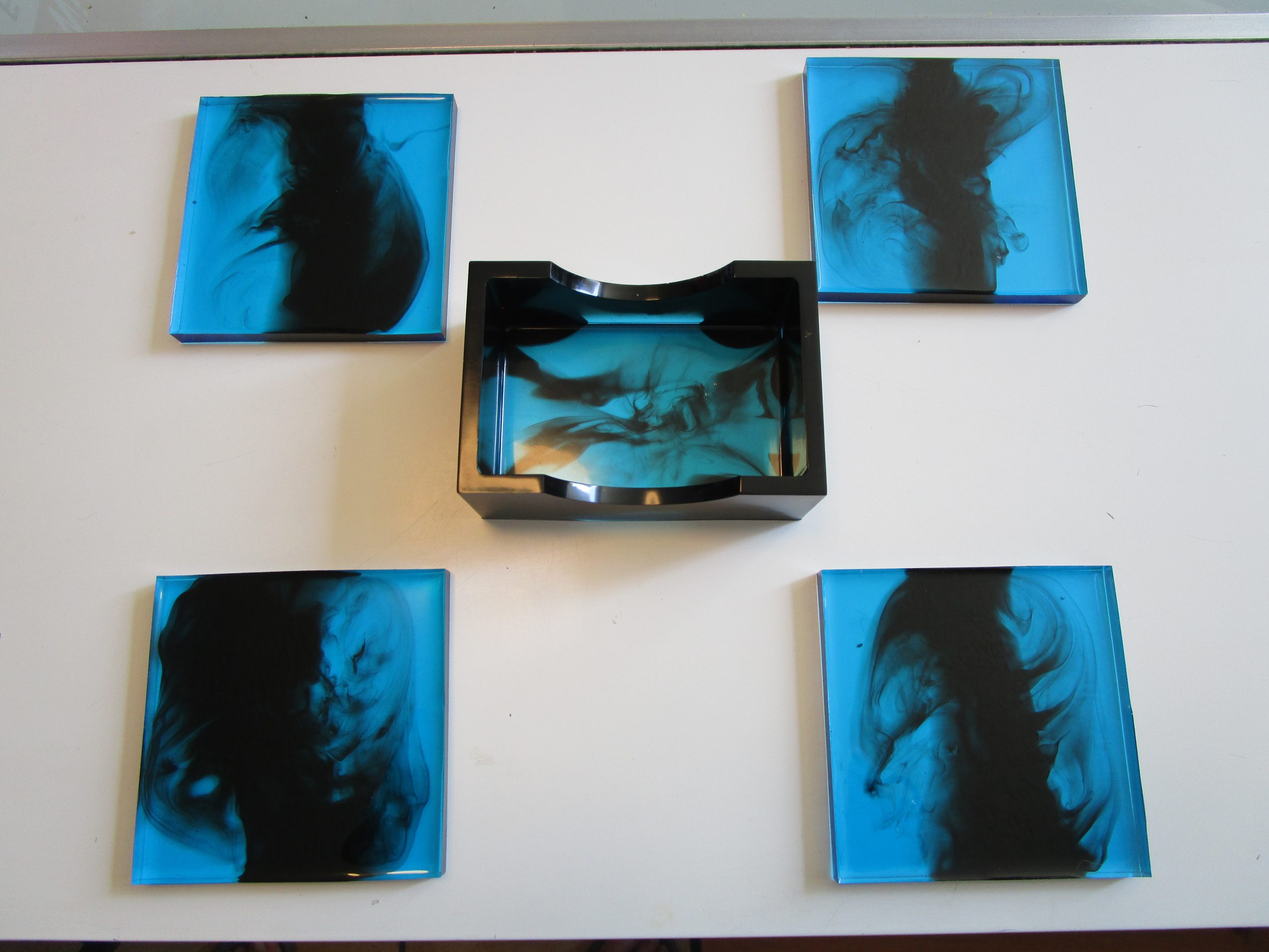 Blue Service. Quatre dessous de verre carrés et un socle de rangement rectangulaire, en résine bleu lagon et noir, posés sur une table blanche.