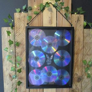 Le Souffle d'un Son. Cadre mural, neuf CDs de musique disposés en forme de lapin sur fond noir. Reflets irisés bleus, violets et verts. Accroché sur un mur en bois avec des guirlandes lumineuses et du lierre.