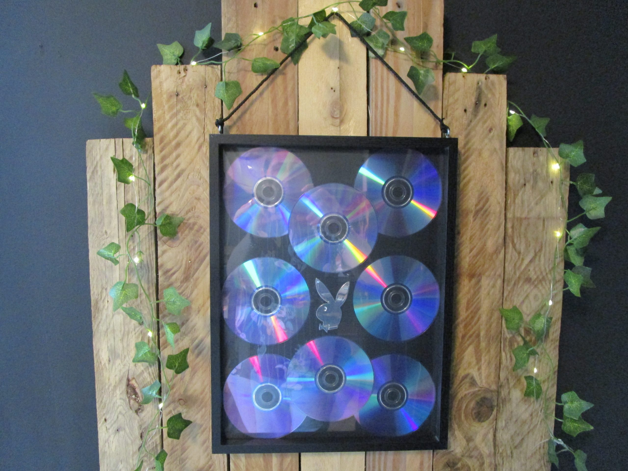 Le Souffle d'un Son. Cadre mural, neuf CDs de musique disposés en forme de lapin sur fond noir. Reflets irisés bleus, violets et verts. Accroché sur un mur en bois avec des guirlandes lumineuses et du lierre.