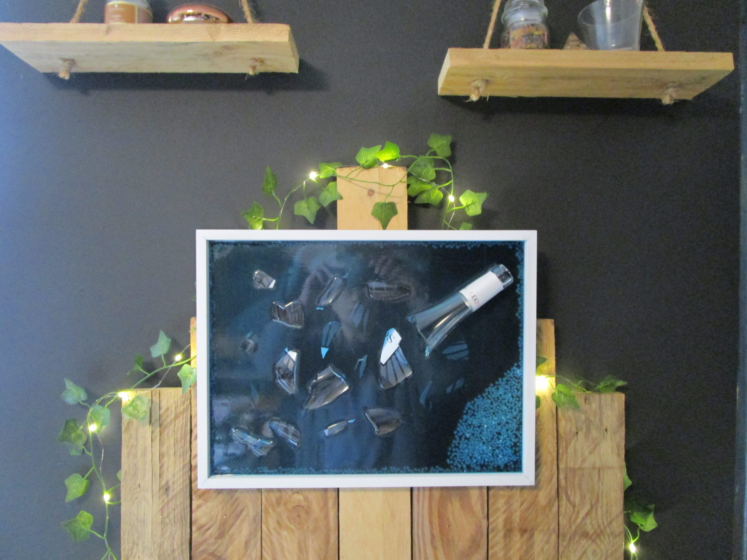 Bouteille à la Mer, fait main en résine époxy bleue foncée, intégrant une bouteille de vin brisée (Léo) et des pierres décoratives sur fond noir, encadré en blanc.