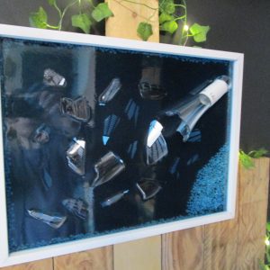 Bouteille à la Mer, fait main en résine époxy bleue foncée, intégrant une bouteille de vin brisée (Léo) et des pierres décoratives sur fond noir, encadré en blanc.