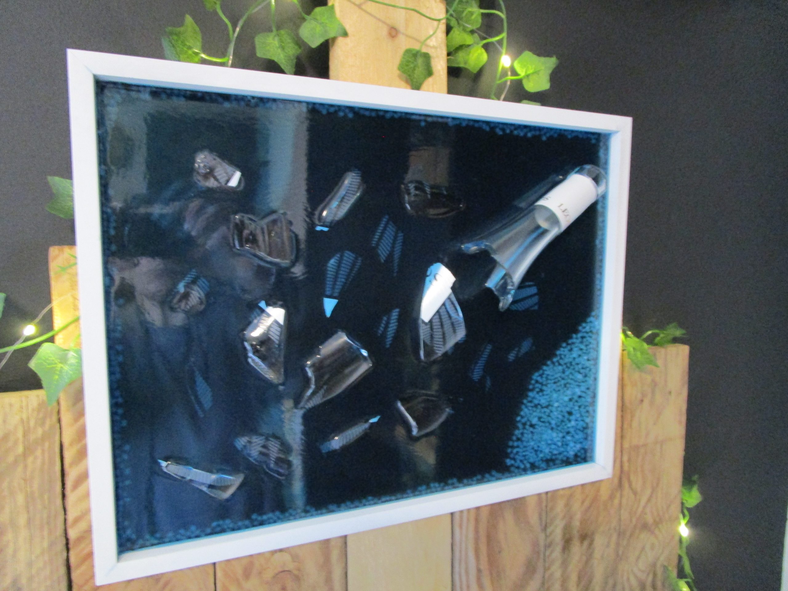 Bouteille à la Mer, fait main en résine époxy bleue foncée, intégrant une bouteille de vin brisée (Léo) et des pierres décoratives sur fond noir, encadré en blanc.