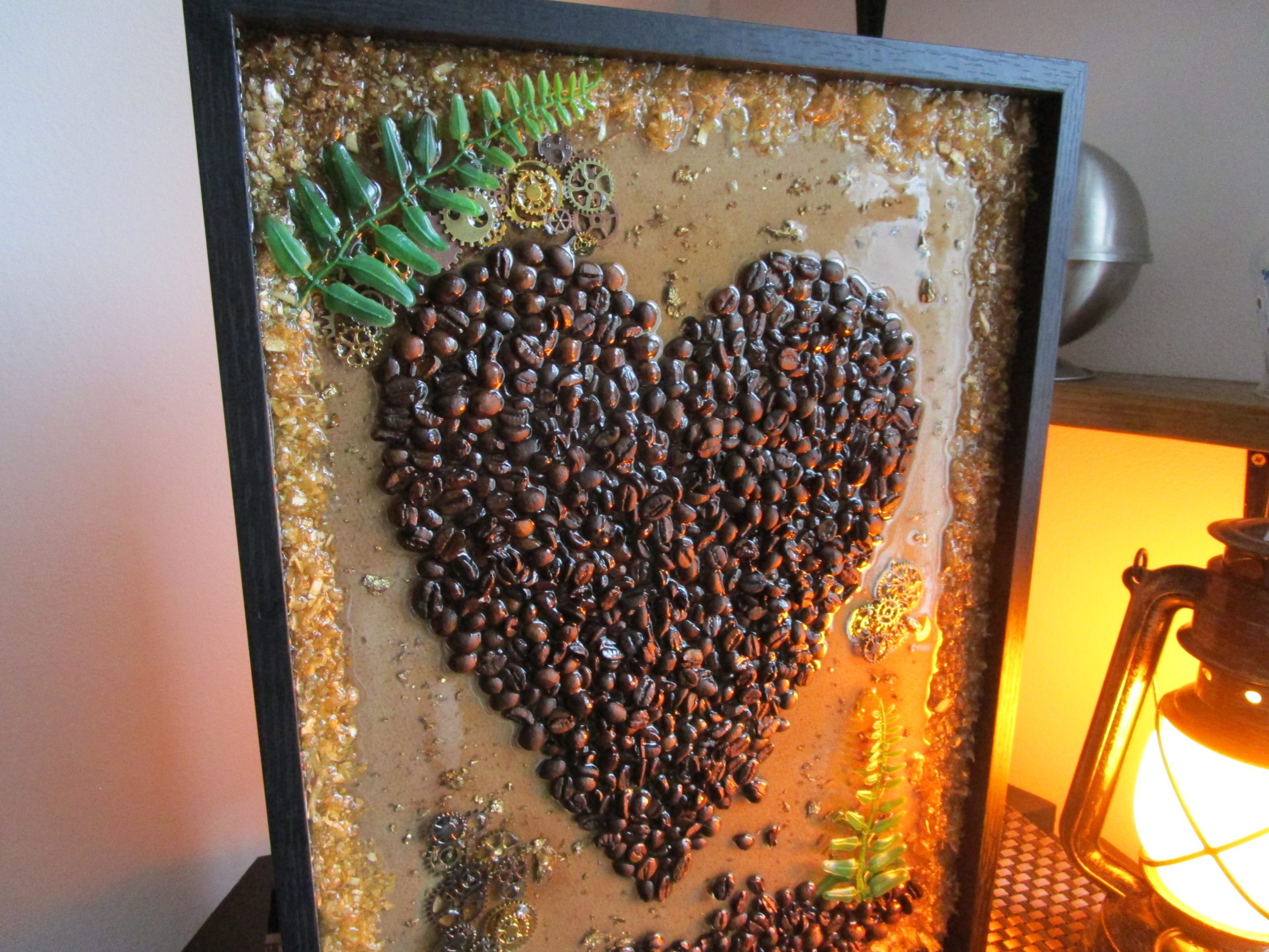 Love Cafée, Cadre décoratif unique Love Cafée en résine avec un cœur central formé de grains de café torréfiés, incrustations de feuilles d'or, de copeaux de bois, de petites feuilles de fougère et de mécanismes d'horlogerie steampunk.