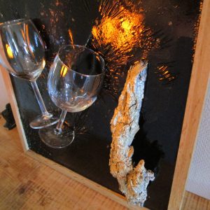 Tableau en résine époxy noir et rouge avec deux verres à vin incrustés, une écorce de bois peinte en doré et des fragments de pierre noire. Cadre en bois clair. Création artisanale "Toast Éternel".