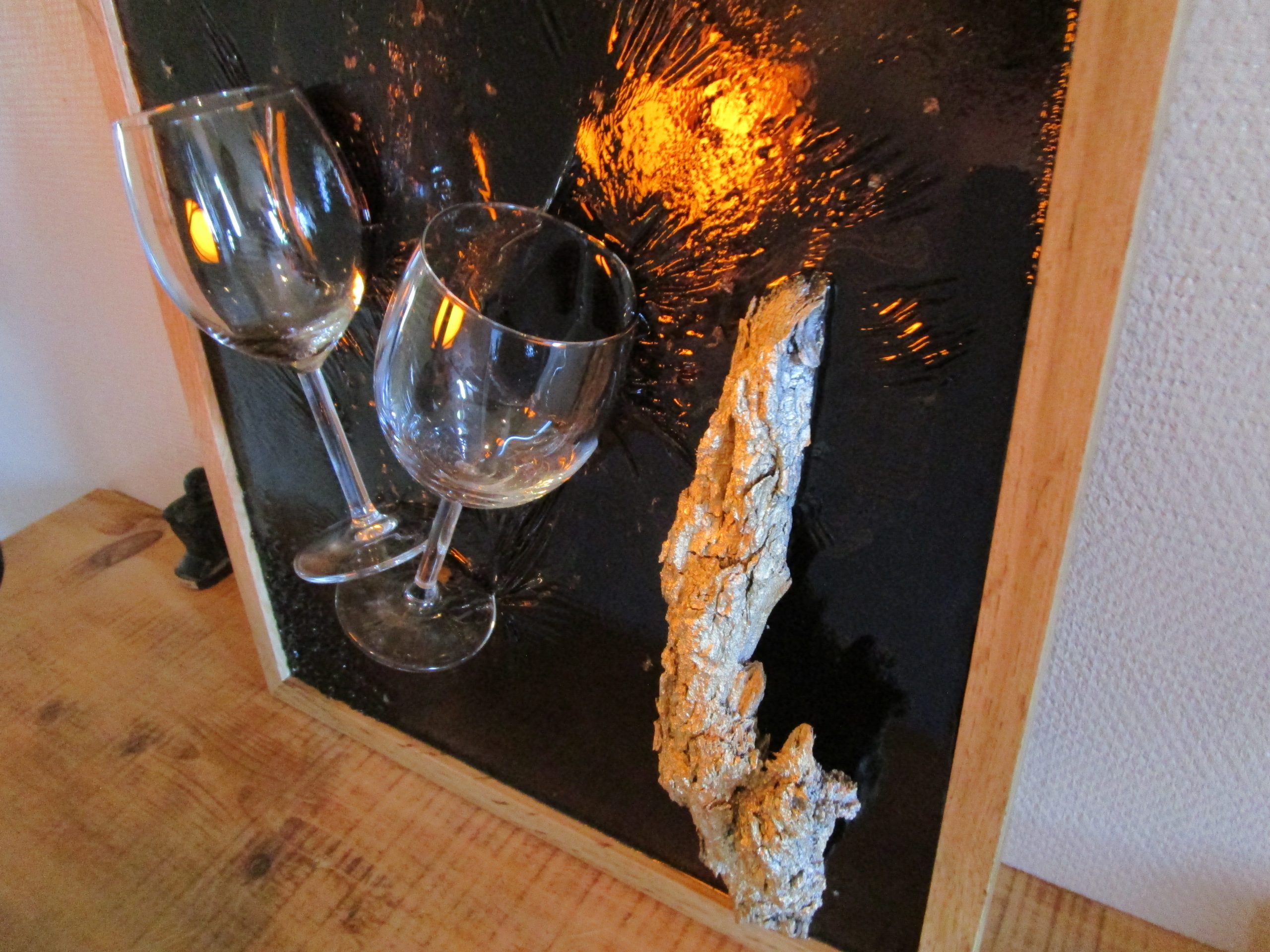 Tableau en résine époxy noir et rouge avec deux verres à vin incrustés, une écorce de bois peinte en doré et des fragments de pierre noire. Cadre en bois clair. Création artisanale "Toast Éternel".