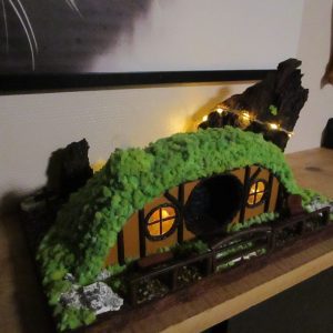 Maison de Hobbit miniature faite main posée sur une étagère en bois. La structure est semi-enterrée sous une colline de mousse verte. La façade montre une porte et deux fenêtres rondes, illuminées par une lumière jaune chaude. Un petit banc est visible à gauche, une chaise à droite, et une barrière en bois délimite le jardin. À l'arrière-plan, un grand morceau d'écorce de bois sombre est éclairé par une fine guirlande lumineuse. Des pierres œil-de-tigre pavent le sol devant l'entrée.