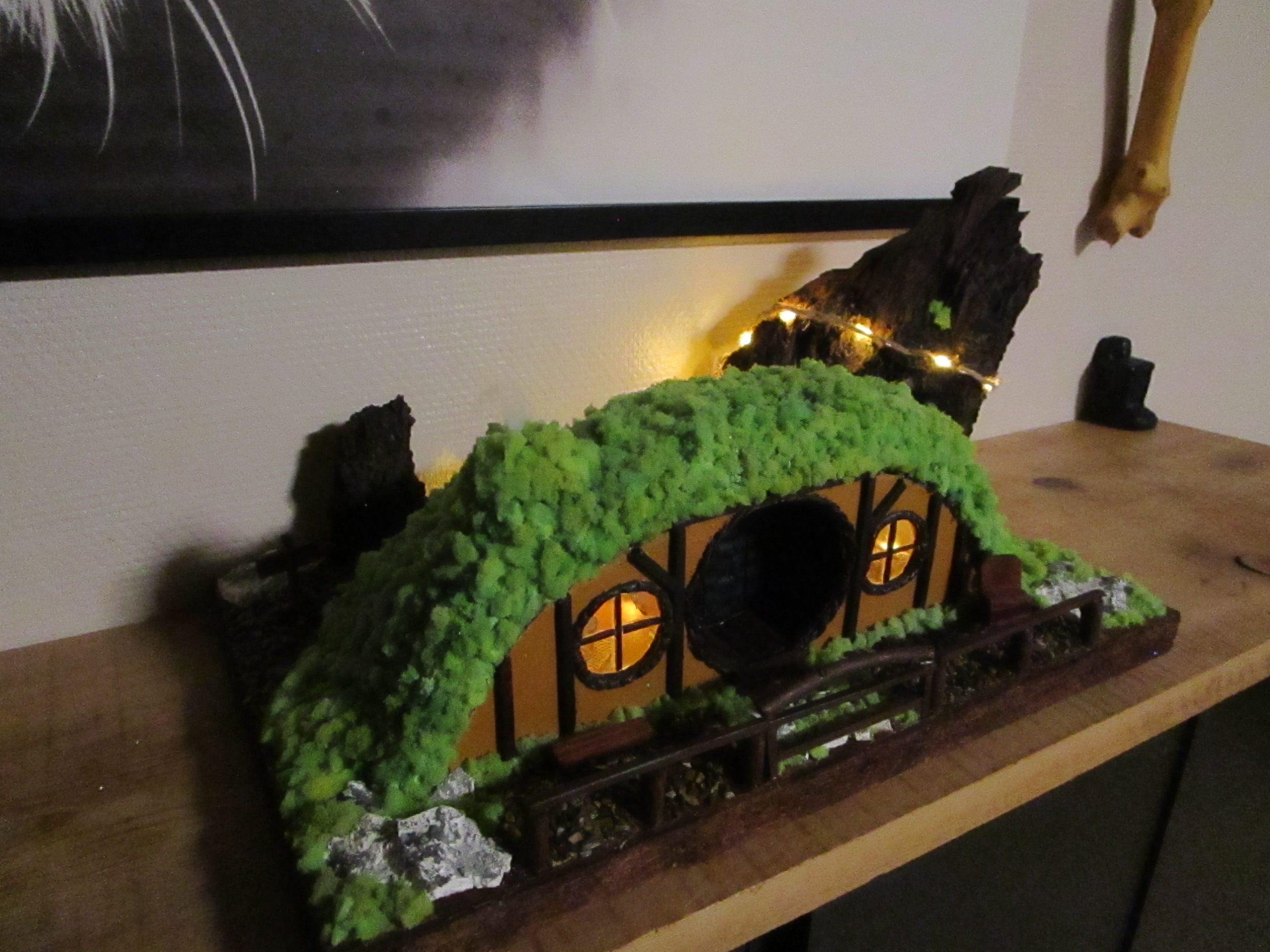 Maison de Hobbit miniature faite main posée sur une étagère en bois. La structure est semi-enterrée sous une colline de mousse verte. La façade montre une porte et deux fenêtres rondes, illuminées par une lumière jaune chaude. Un petit banc est visible à gauche, une chaise à droite, et une barrière en bois délimite le jardin. À l'arrière-plan, un grand morceau d'écorce de bois sombre est éclairé par une fine guirlande lumineuse. Des pierres œil-de-tigre pavent le sol devant l'entrée.