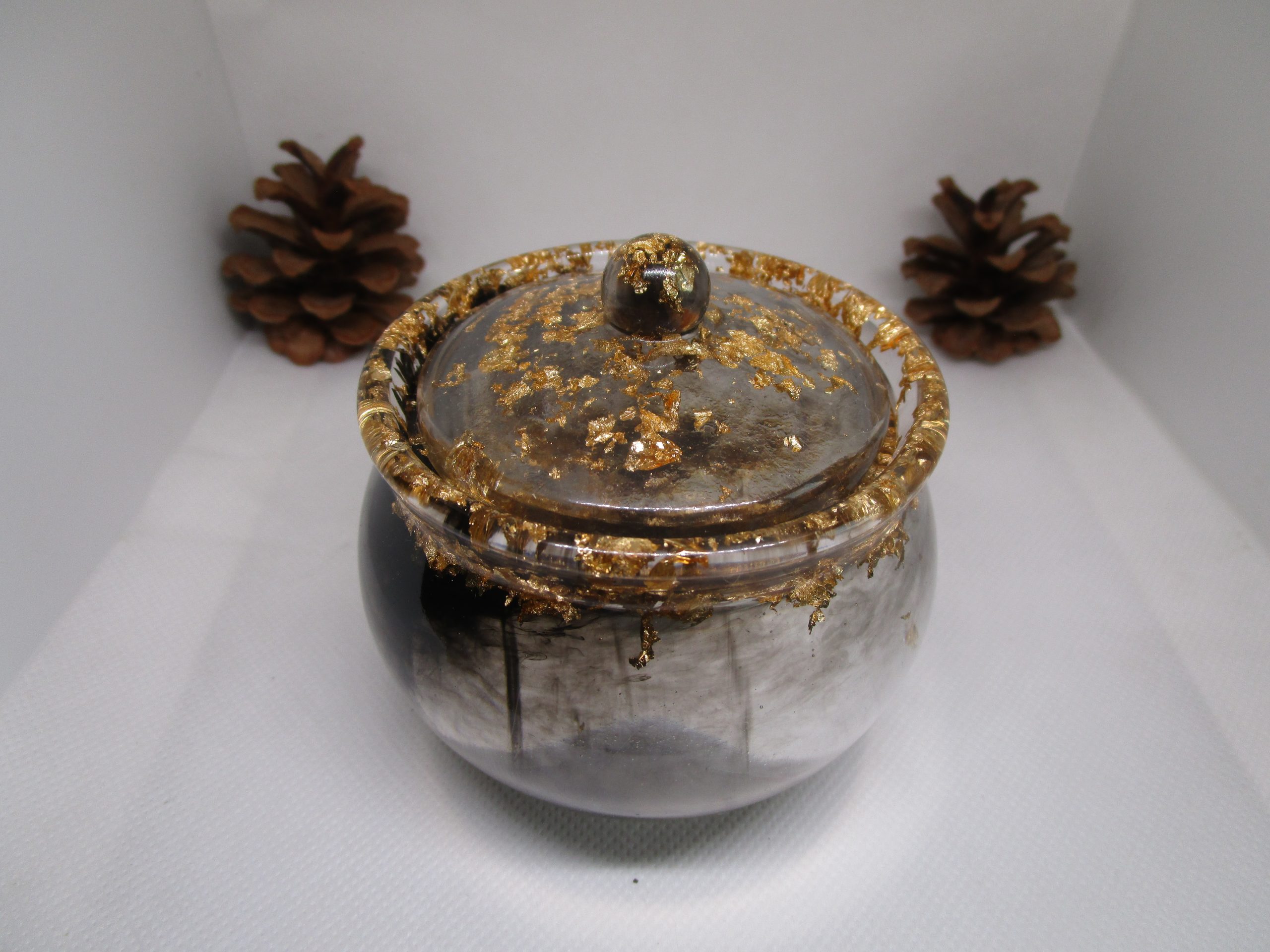 Pot de rangement fait main en résine époxy noire et transparente avec couvercle, incrusté de feuilles d'or autour du bord, posé à côté d'une pomme de pin.