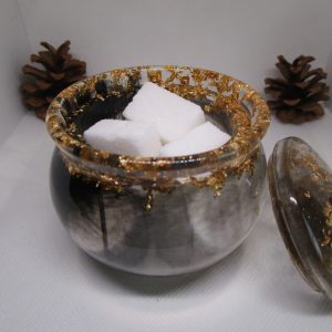 Pot de rangement fait main en résine époxy noire et transparente avec couvercle, incrusté de feuilles d'or autour du bord, posé à côté d'une pomme de pin.