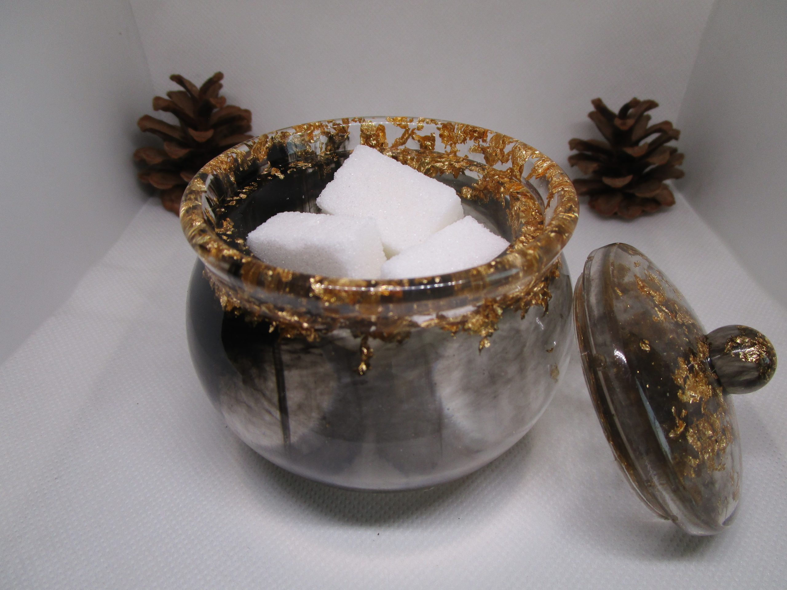 Pot de rangement fait main en résine époxy noire et transparente avec couvercle, incrusté de feuilles d'or autour du bord, posé à côté d'une pomme de pin.