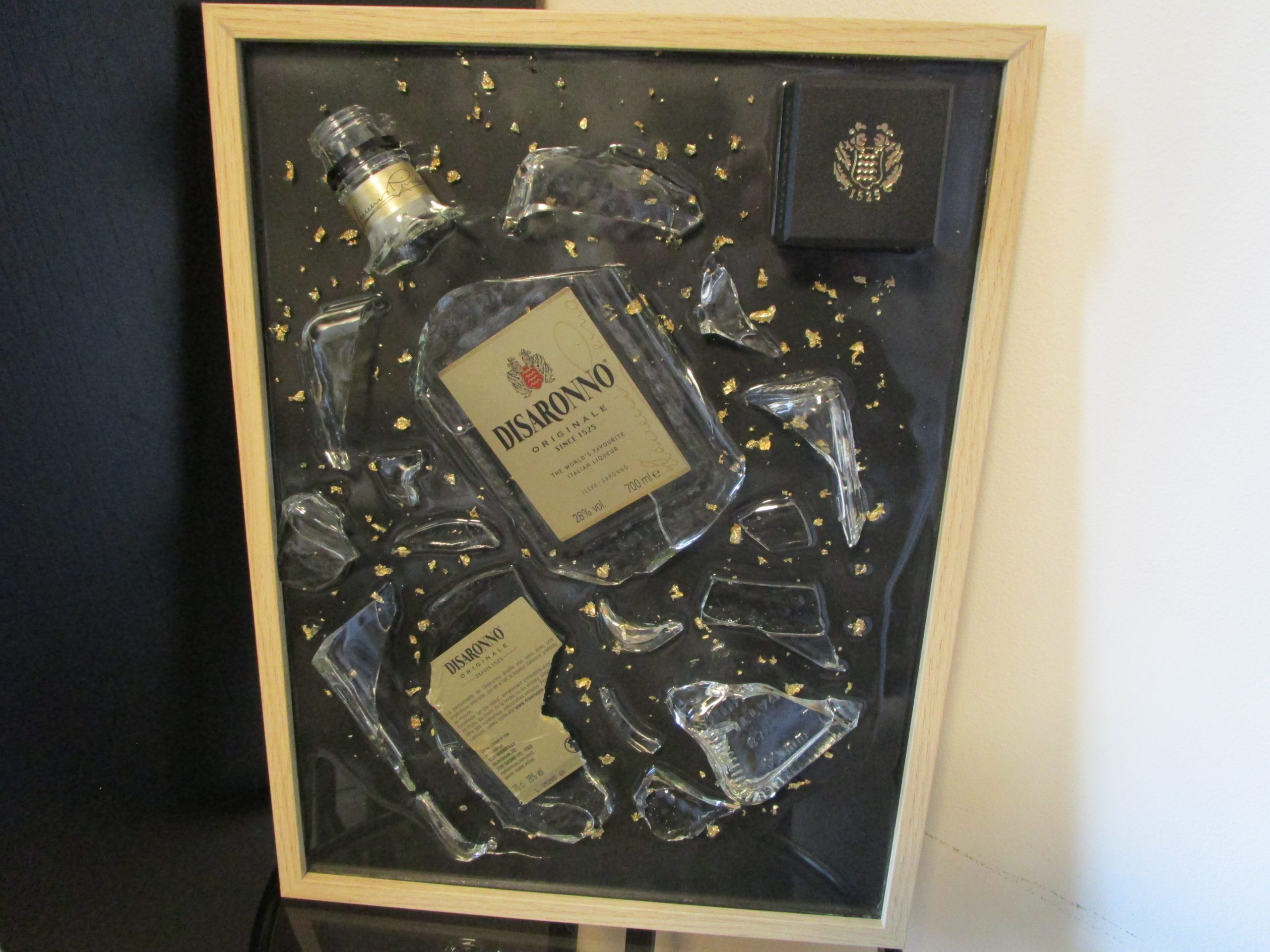 Tableau en résine d'artisanat d'une bouteille d'Amaretto Disaronno brisée, avec des éclats de verre et des fragments de feuilles d'or sur fond noir, encadré de bois clair.