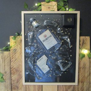 Tableau en résine d'artisanat d'une bouteille d'Amaretto Disaronno brisée, avec des éclats de verre et des fragments de feuilles d'or sur fond noir, encadré de bois clair.