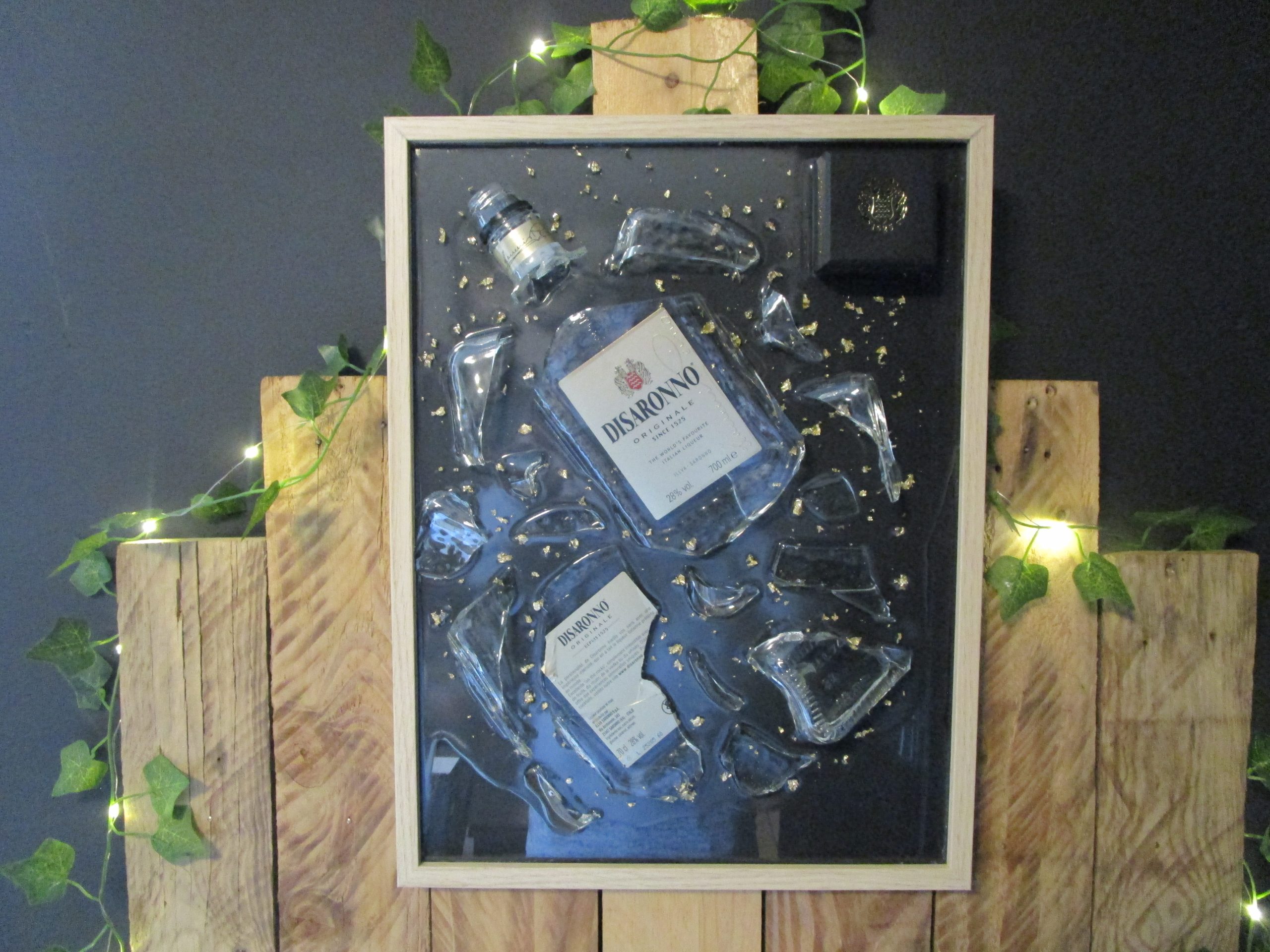 Tableau en résine d'artisanat d'une bouteille d'Amaretto Disaronno brisée, avec des éclats de verre et des fragments de feuilles d'or sur fond noir, encadré de bois clair.