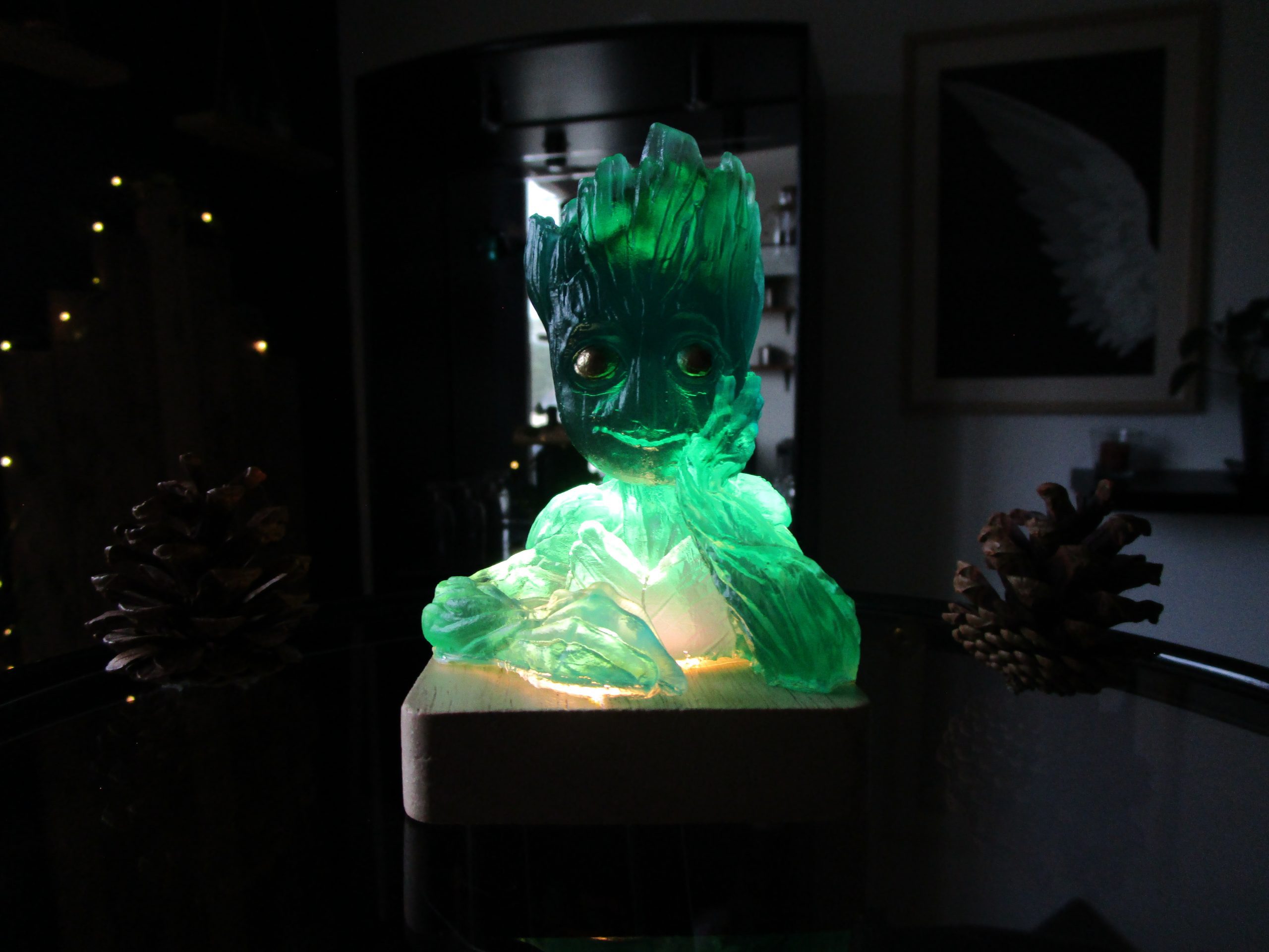 Figurine artisanale Le Groot des Rivières en résine translucide vert émeraude et bleu lagon, posée sur un socle en bois clair illuminé par l'intérieur. La lumière met en valeur l'aspect cristallin du buste de Groot dans une ambiance sombre avec des pommes de pin décoratives.