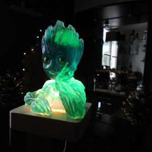 Figurine artisanale Le Groot des Rivières en résine translucide vert émeraude et bleu lagon, posée sur un socle en bois clair illuminé par l'intérieur. La lumière met en valeur l'aspect cristallin du buste de Groot dans une ambiance sombre avec des pommes de pin décoratives.