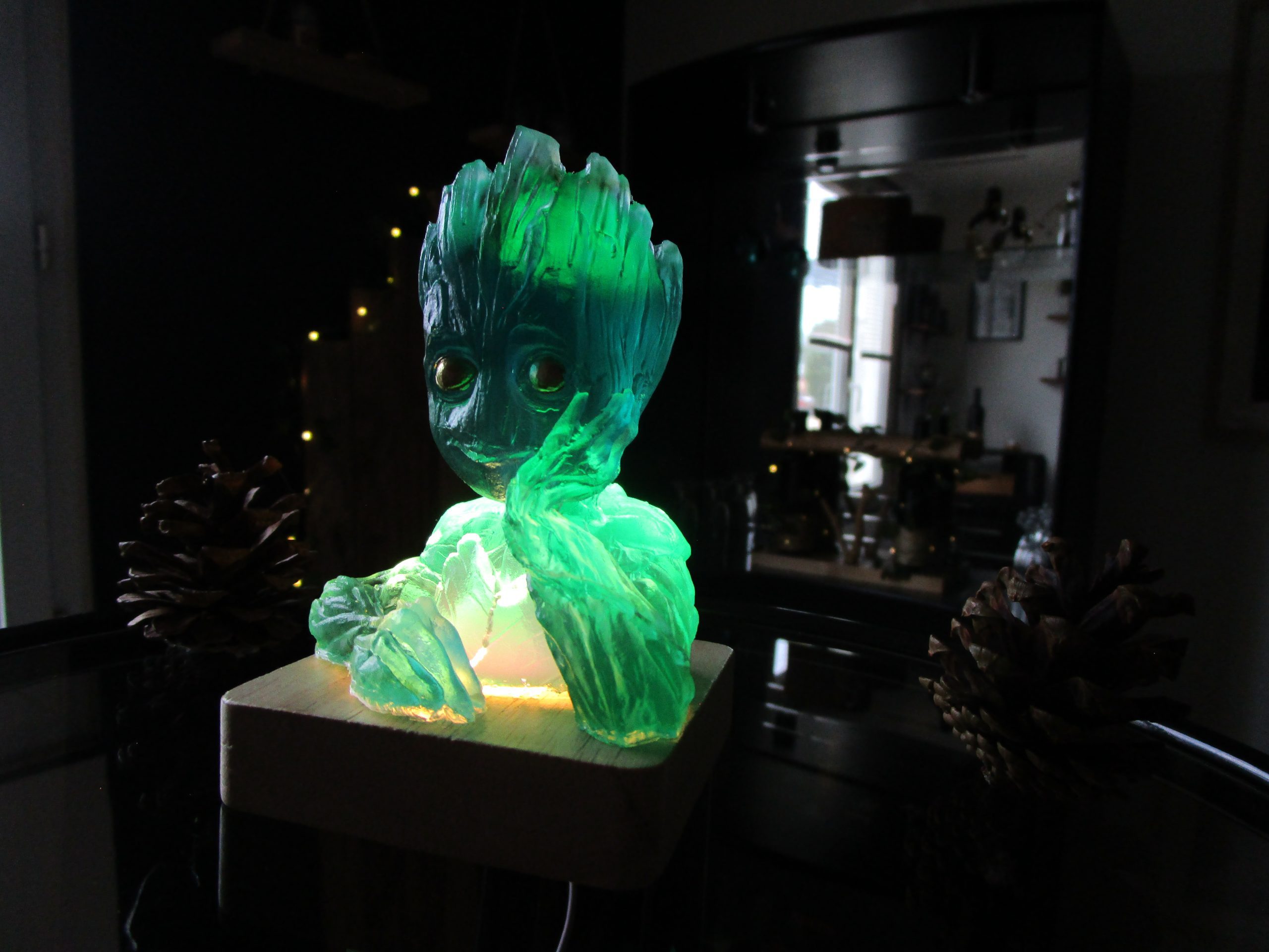Figurine artisanale Le Groot des Rivières en résine translucide vert émeraude et bleu lagon, posée sur un socle en bois clair illuminé par l'intérieur. La lumière met en valeur l'aspect cristallin du buste de Groot dans une ambiance sombre avec des pommes de pin décoratives.