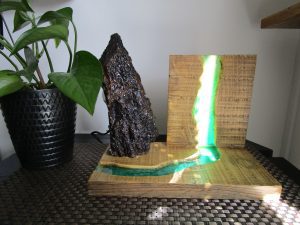 Lampe artisanale en bois massif avec une inclusion de résine époxy turquoise translucide créant l'effet d'une rivière rétroéclairée, surmontée d'une écorce de bois brut texturée.