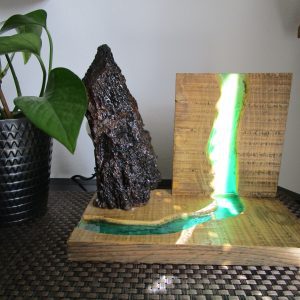 Lampe artisanale en bois massif avec une inclusion de résine époxy turquoise translucide créant l'effet d'une rivière rétroéclairée, surmontée d'une écorce de bois brut texturée.