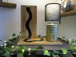 Création artisanale composée d'une planche en bois lasuré intégrant une rivière sculptée en résine noire brillante, accompagnée d'une lampe cylindrique dorée avec abat-jour noir.